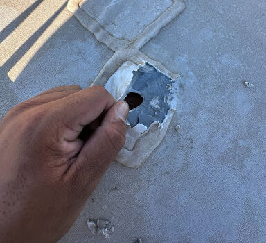 A hand peels back cracked caulking from a rooftop patch around a former antenna base, exposing underlying damage—highlighting why relying on caulk alone for roof repairs can lead to long-term issues.